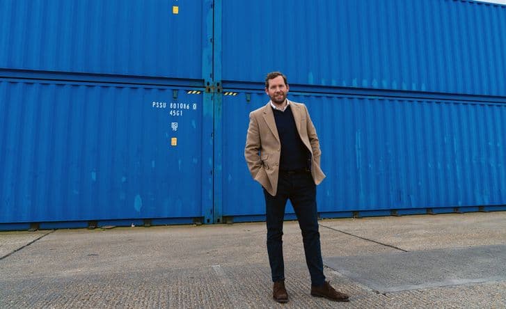 A photo of Johnny McCord in a light brown jacket, black jumper and white shirt, standing with his hands in his pockets in front of several blue shipping containers.