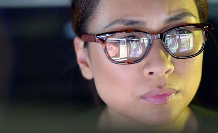 A close-up of a woman's face, she is wearing round brown glasses. You can see the reflection of two screens in her glasses.