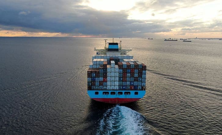 An image from behind and above of a cargo ship out at sea, it's heavily loaded with many shipping containers. There are other ships in the background too.