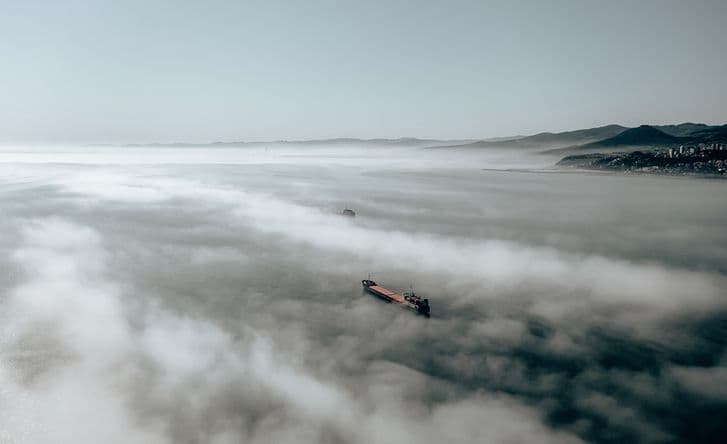 A landscape photo of the sea, which is almost completely covered by fog. You can see the coastline on the right and one cargo boat emerging through the mist.