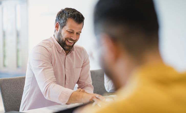A man in a pink shirt is smiling, he is sitting at a table in a meeting with some other people, but they are blurred.