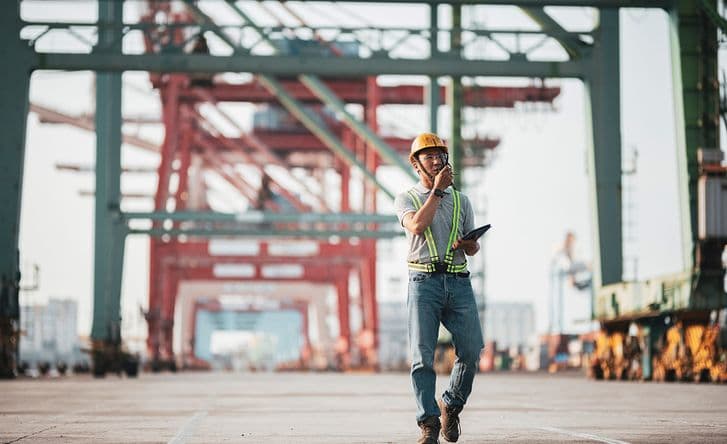 A man in an industrial setting is wearing a hard hat and speaking to someone on a radio.