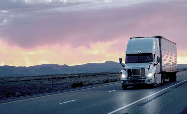 An image of a white cargo truck driving alone a highway with pink clouds behind it, headlights on.