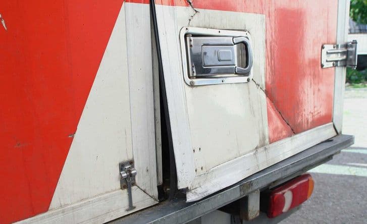 A photo showing the corner of a red and white cargo truck where it's clear that entry has been forced - the door is bent and a crack runs through the cover.