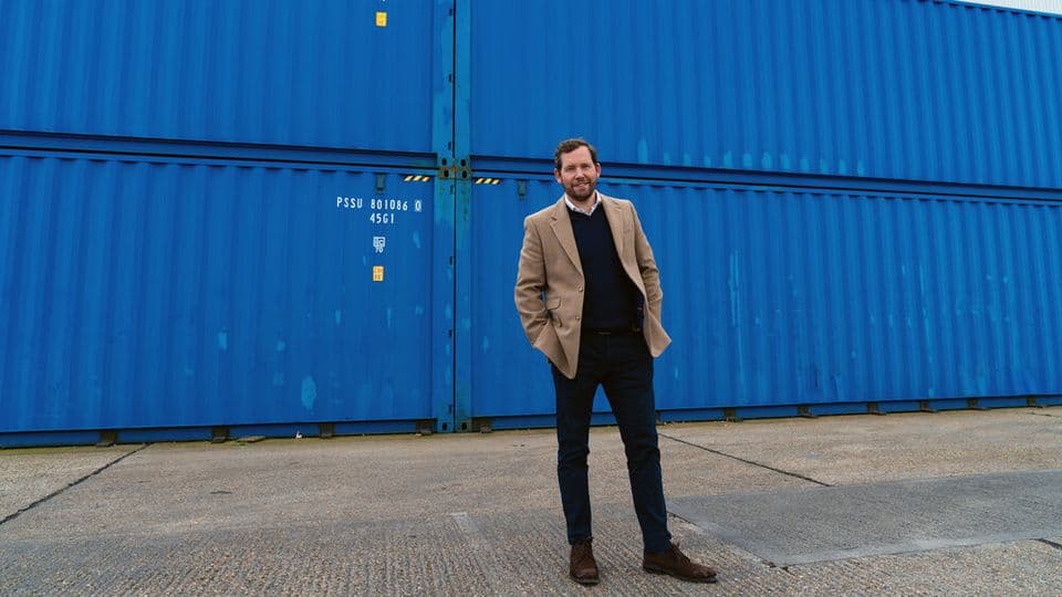 A photo of Johnny McCord in a light brown jacket, black jumper and white shirt, standing with his hands in his pockets in front of several blue shipping containers.