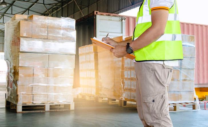 A shot from the shoulders down of a man standing in a warehouse full of wrapped pallets. He is wearing high vis and holding an orange clipboard.