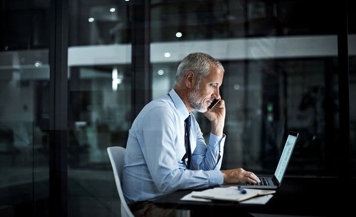 A man sits in an office wearing a suit, he's on the phone and using his laptop at the same time.