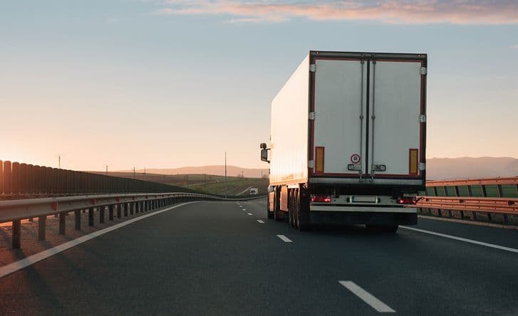 A white cargo truck is pictured from behind as it drives along the right lane of a highway. The sun is setting on the left.