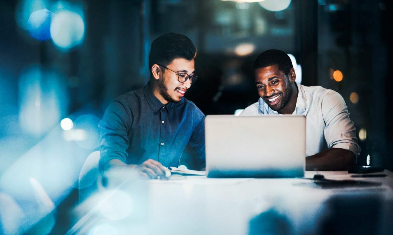 Two men are sitting next to each other looking at a laptop and smiling.