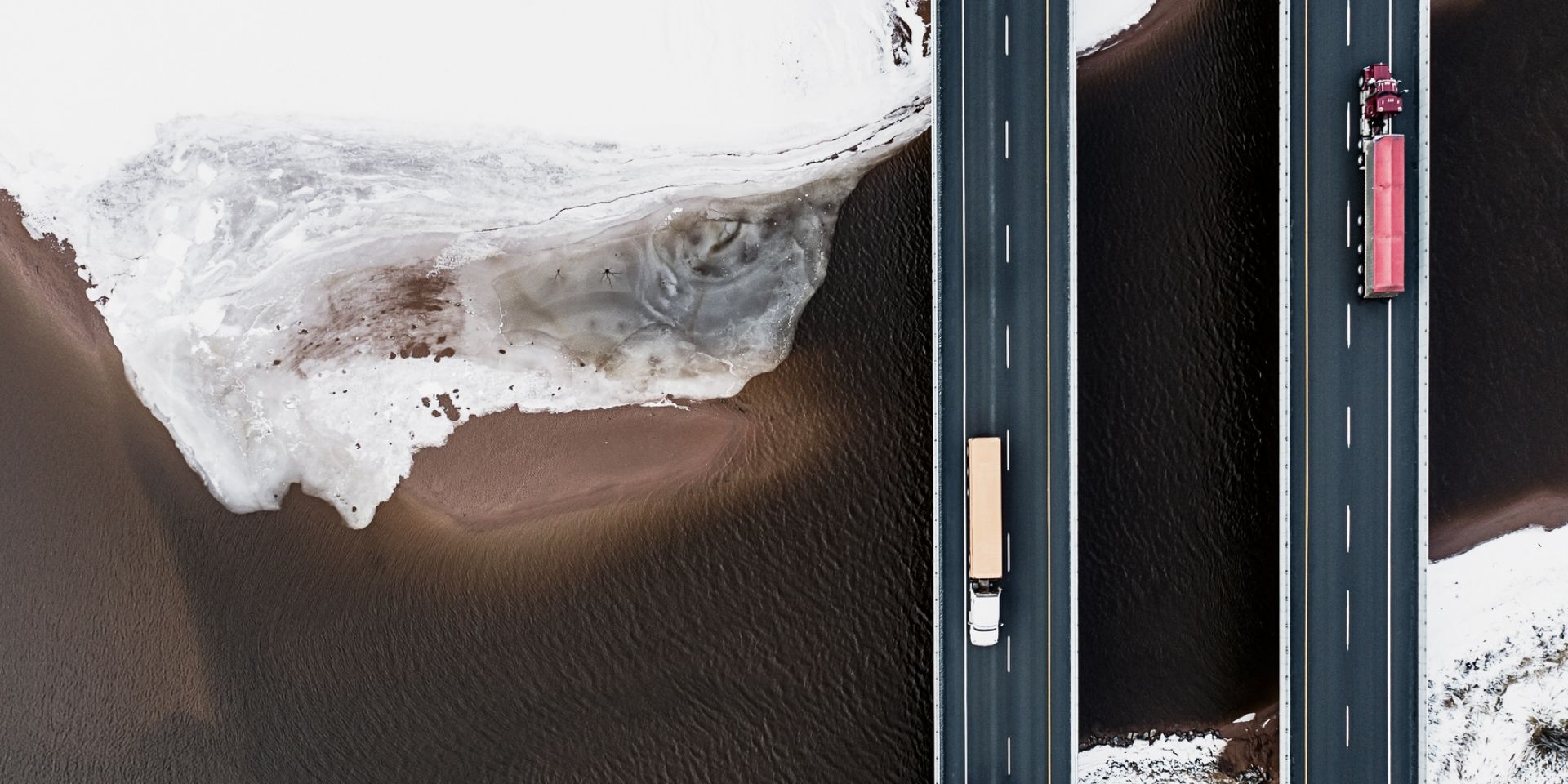 An image of two highways bridging over a brown river bed, with a red truck going in one direction and yellow truck in the other.