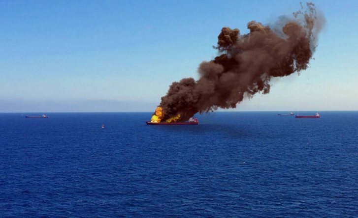 A landscape of the sea with a marine cargo boat in the middle, which is on fire. Large yellow flames send black clouds of smoke up into the air.