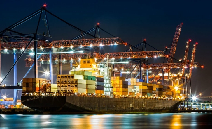A photo of a marine cargo ship lit up at night, while it docks. There are several cranes operating and the ship yard is lit up.
