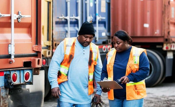 A photo of two people in a shipyard looking at a clipboard, both wearing orange high vis.