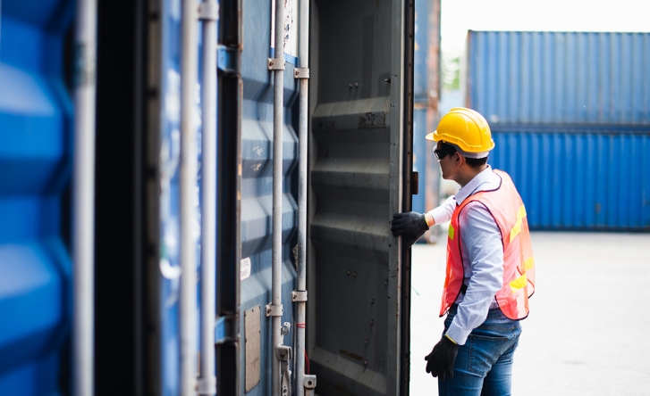 A man in protective gear is opening a shipping container door in a ship yard. There are blue shipping containers in the background. =