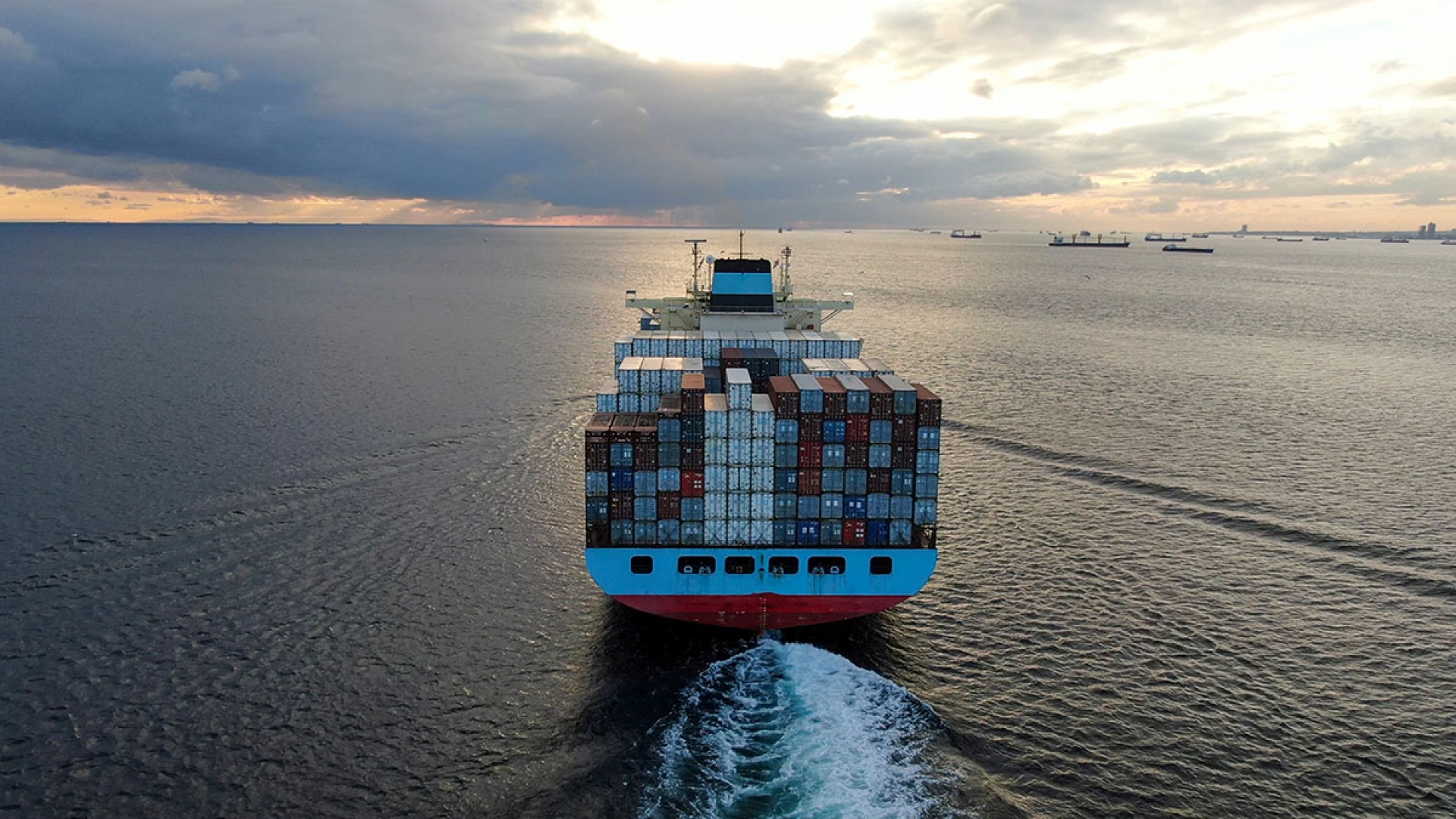 An image from behind and above of a cargo ship out at sea, it's heavily loaded with many shipping containers. There are other ships in the background too.