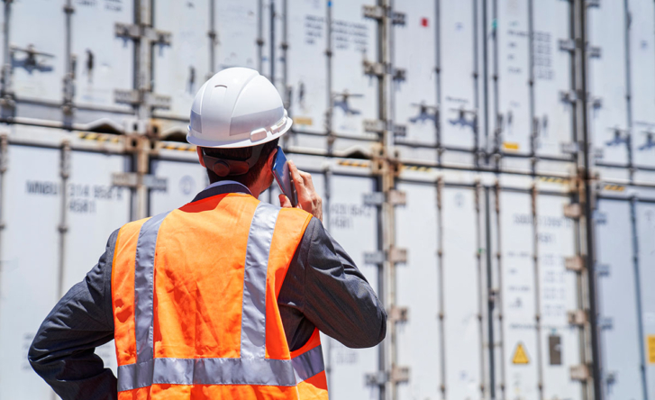A man in high vis and a hard hat is standing in front of a pile of shipping containers, on the phone. We see him from behind.