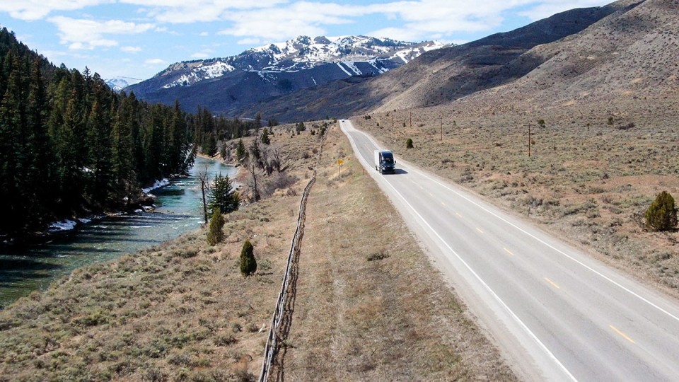 An image of a cargo truck driving along a highway past a forest and a river, with mountains in the background.