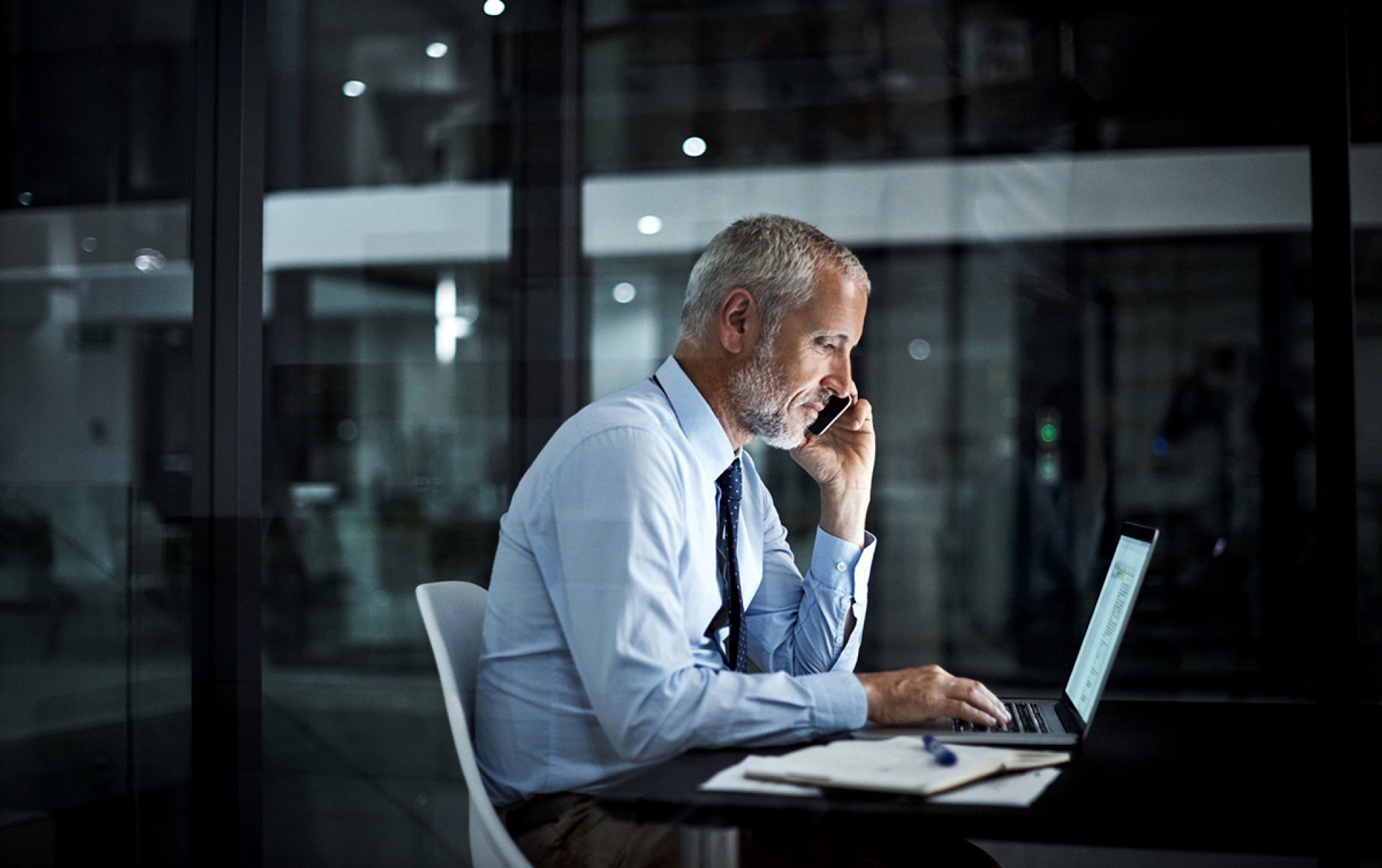 A man sits in an office wearing a suit, he's on the phone and using his laptop at the same time.