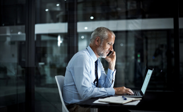 A man sits in an office wearing a suit, he's on the phone and using his laptop at the same time.
