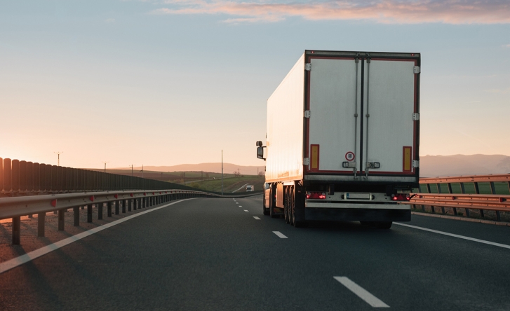 A white cargo truck is pictured from behind as it drives along the right lane of a highway. The sun is setting on the left.