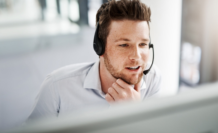 A man with brown hair is sitting at a desk speaking into a headset, he is smiling and wearing an off-shite shirt.