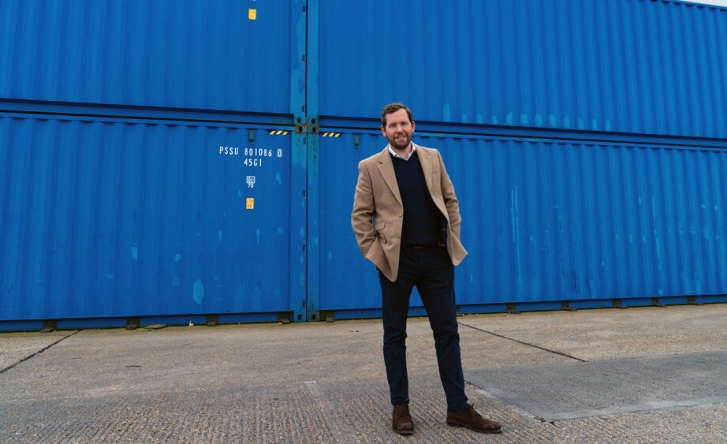 A photo of Johnny McCord in a light brown jacket, black jumper and white shirt, standing with his hands in his pockets in front of several blue shipping containers.