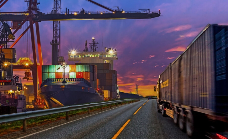 A black cargo truck driving down a highway at sunset, past a lit up ship yard.