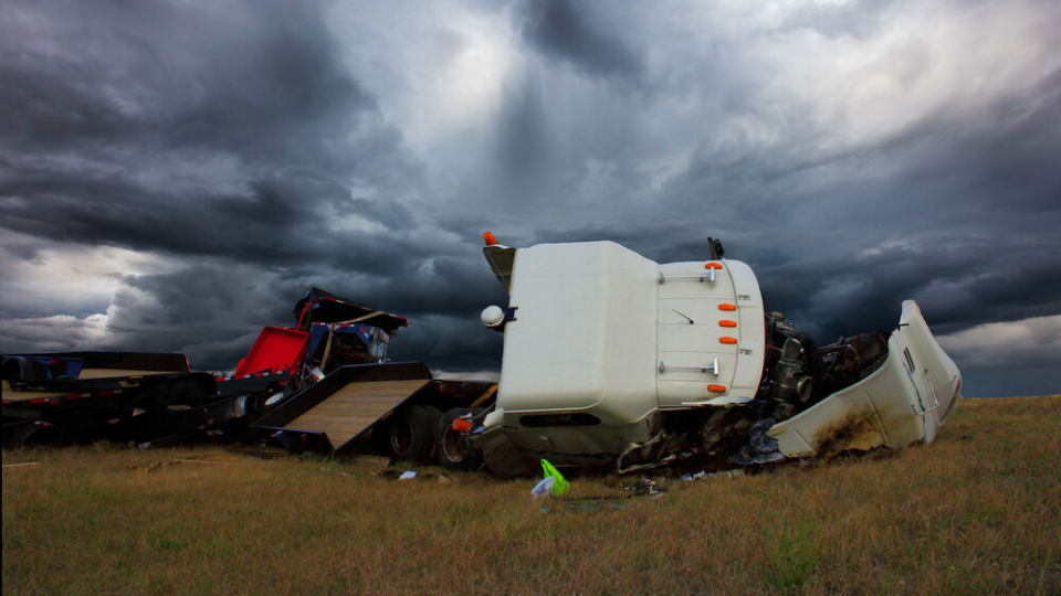 A photo of the wreckage of a white cargo truck on its side, with a stormy sky behind it.