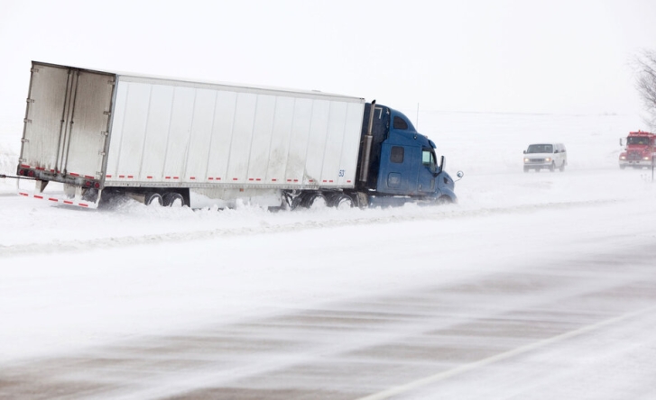 A cargo truck that has come off the road in the snow, with a blue cabin. Two cars are approaching it from the other direction.