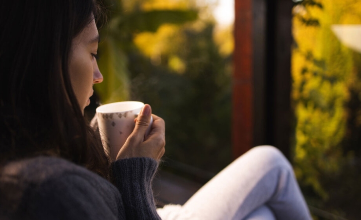 A close up of a woman holding a hot drink in a mug, with her legs crossed as she looks out of the window.